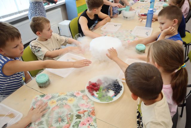 Na zdjęciu znajdują się dzieci przy złączonych stolikach i wyciągają ręce do miski, z której unosi się dym od suchego lodu, na stoliku leżą również serwetki, tacki.