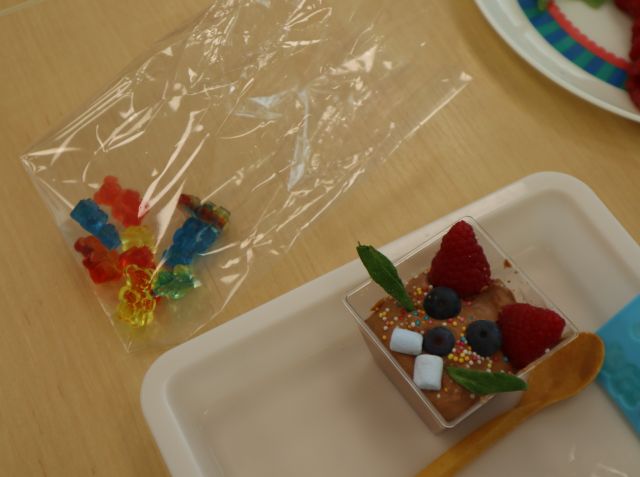 Na zdjęciu znajdują się kolorowe żelki w plastikowej foli oraz ozdobiony mus czekoladowy, łyżeczka obok.