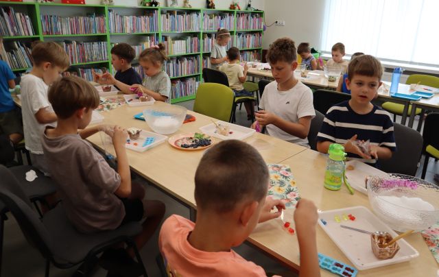 Na zdjęciu znajdują się dzieci przy złączonych w dwóch rzędach stołach i wyciągają swoje żelki z foremek. Na stoliku leżą miski, miseczki, tacki, owoce, a w tle znajdują się półki z książkami.