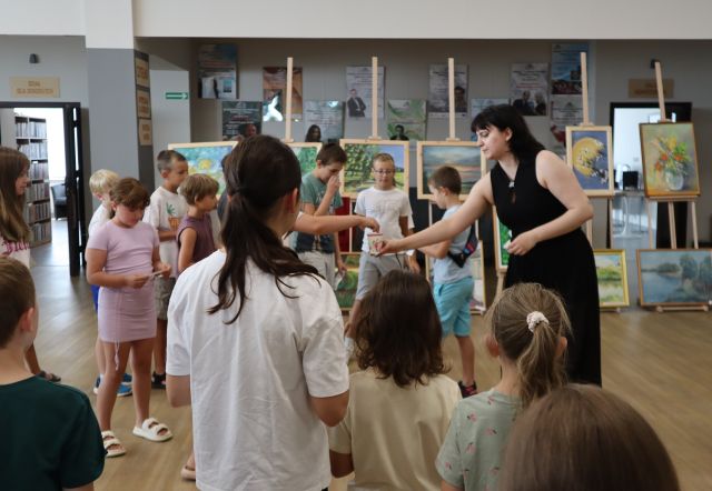 Na zdjęciu znajduje się grupka dzieci wraz z kobietą, która daje im do losowania karteczki. Warsztaty odbywają się na holu biblioteki, a w tle znajdują się obrazy oraz plakaty.