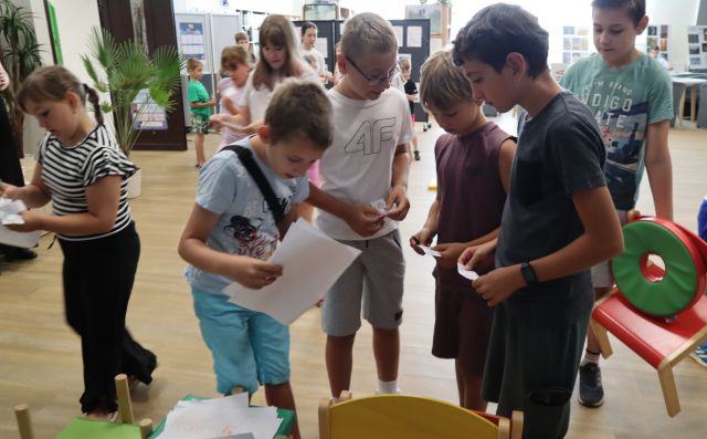 Na zdjęciu znajdują się dzieci, w centrum znajduje się pięciu chłopców, którzy trzymają w ręce kartki, trzech z nich trzyma małe karteczki,a jeden dużą, jeden z chłopców stojący za nimi trzyma krzesełko oraz pufę. W tle znajdują się plakaty oraz czytelnia.