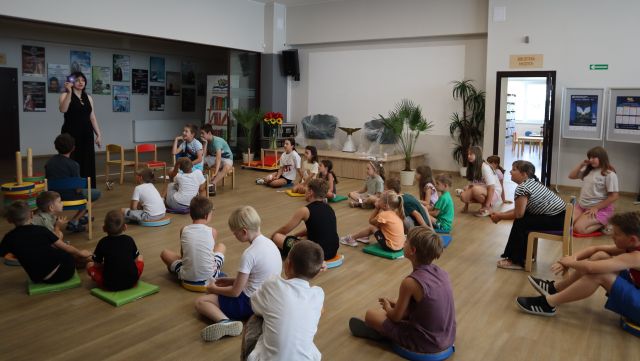 Na zdjęciu znajdują się dzieci siedzące na pufach na holu biblioteki, po lewej stronie stoi kobieta i pokazuje karteczkę uczestnikom zajęć, obok niej stoją krzesełka. W tle znajdują się drzwi, scena z krzesłami, a obok niej rośliny w doniczkach, plakaty na ścianach.