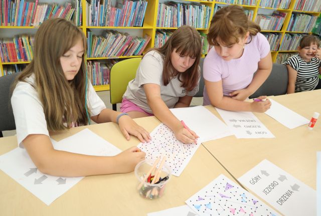 Na zdjęciu znajdują się cztery dziewczynki siedzące przy złączonych stołach, trzy z nich wspólnie rozwiązują zadania na kartkach przed sobą, na środku leżą również kredki. W tle znajdują się półki z książkami.