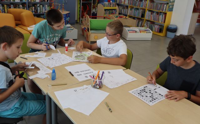 Na zdjęciu znajduje się czwórka chłopców przy złaczonych stolikach, którzy rozwiązują zadania z kodowania na kartkach przed sobą, na środku znajdują się również kredki w pojemniku i klej. W tle znajdują się półki z książkami, samochód do zabawy oraz okrągła pufa.