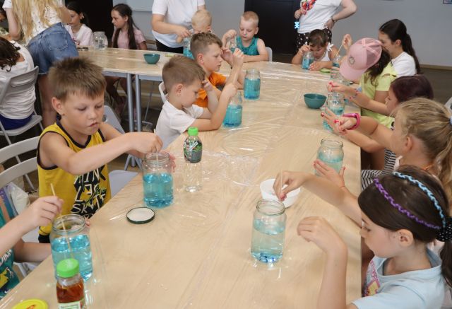 Na zdjęciu znajdują się dzieci przy złączonych stołach owiniętych folią i mieszają wodę z niebieskim barwnikiem w słoikach.