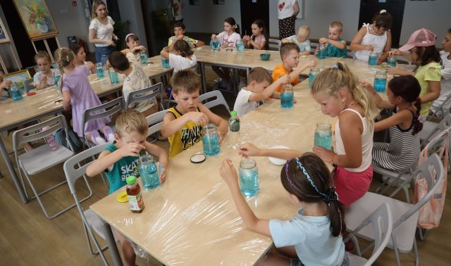 Na zdjęciu znajduje się około 20 dzieci przy złączonych w podkowę stołach, uczestnicy mieszają niebieski barwnik w słoikach z wodą. Przy stołach stoją dwie kobiety, a w tle znajduje się obraz.