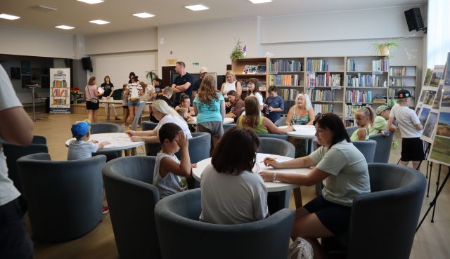 Na zdjęciu znajdują sie cztery okrągłe stoliki, przy których na fotelach siedzą rodzice z dziećmi. W tle znajdują się półki z książkami oraz hol biblioteki.