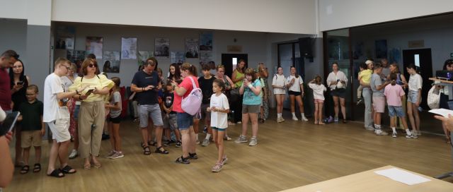 Na zdjęciu znajdują się dorośli i dzieci z telefonami, w tle znajdują się plakaty na ścianach.