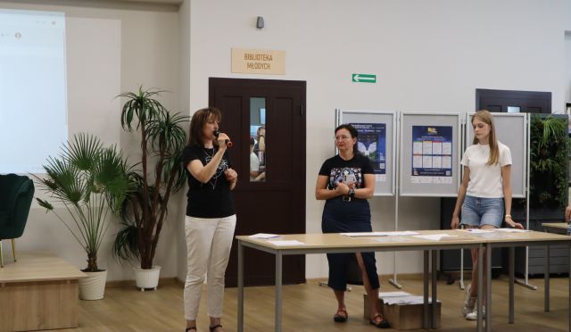 Na zdjęciu znajdują się trzy kobiety przy złączonych stolikach, na których leżą kartki. Kobieta po lewej mówi przez mikrofon. W tle znajdują się dwie rośliny, tablice z plakatami oraz drzwi do Biblioteki Młodych.