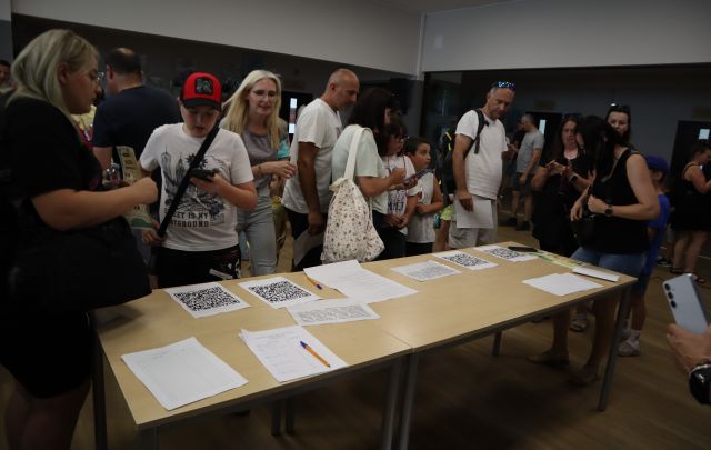 Na zdjęciu znajdują się uczestnicy gry przy złączonych stolikach, na których znajdują się kartki oraz kody QR do gry.