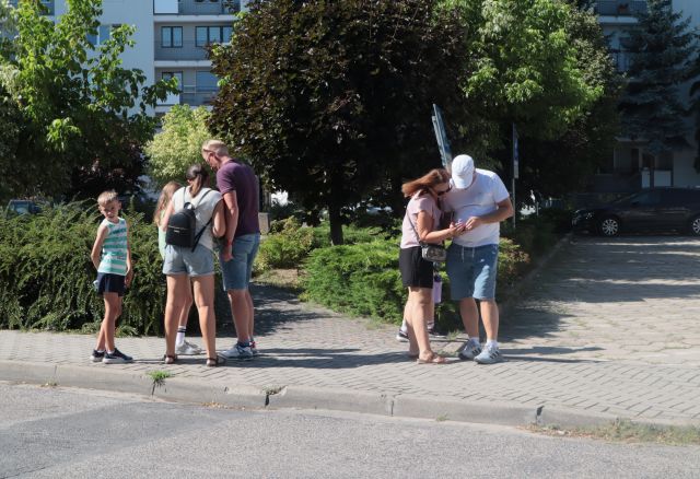 Na zdjęciu znajdują się dwie grupy, jedna dwuosobowa, druga czteroosobowa i rozwiązują zadania z gry miejskiej. W tle znajdują się krzaki, drzewa oraz bloki mieszkalne.
