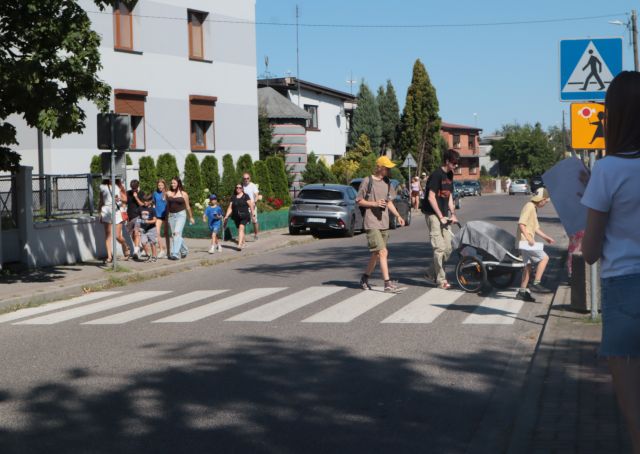 Na zdjęciu znajdują się grupy biorące udział w grze Wawrzynkowej, kilka grup idzie po chodniku po lewej stronie, a część przechodzi na pasach na prawą. W tle znajduje się ulica z domami mieszkalnymi.