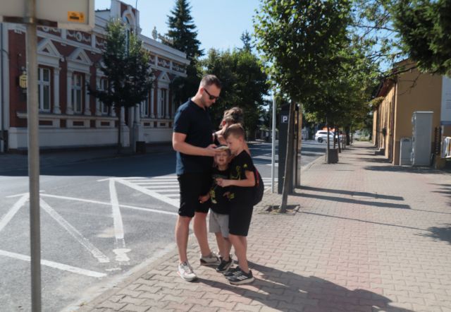 Na zdjęciu znajduje się dwójka chłopców z rodzicami i rozwiązują zadanie na smartfonie z gry miejskiej. Rodzina znajduje się na chodniku, po lewej znajduje się jezdnia oraz po obu stronach znajdują się budynki.