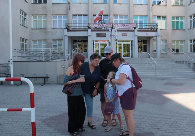 Na zdjęciu znajduje się czwórka dorosłych oraz jedna dziewczynka i rozwiązują zadanie na smartfonie na tle Szkoły Podstawowej.