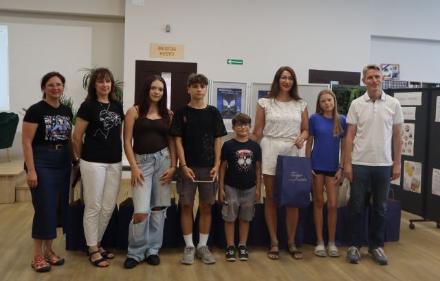Na zdjęciu znajdują się trzy kobiety, jeden mężczyzna, trójka młodzieży oraz chłopiec, którzy stoją do wspólnego zdjęcia jedna z kobiet trzyma grantową torbę. W tle znajdują się tablice z plakatami, drzwi.