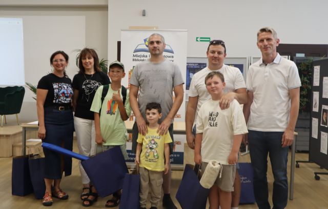 Na zdjęciu znajdują się dwie kobiety, trójka mężczyzn oraz trzech chłopców, którzy trzymają granatowe torby z upominkami. W tle znajduje się baner z logiem biblioteki oraz tablice z plakatami.