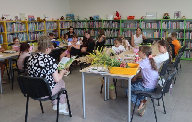 Na zdjęciu znajdują się dwa rzędy zdłączonych stolików przy których siedzą dzieci i dwie kobiety, na środku przed stolikami siedzi kobieta i czyta książkę. W tle znajdują się półki z książkami.