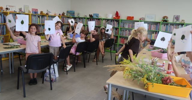 Na zdjęciu znajdują się dzieci przy stolikach w dwóch rzędach, część z nich stoi, część siedzi i pokazują do zdjęcia swoje prace z liśćmi. Na stolikach w pudełkach leżą liście. W tle znajdują się półki z książkami.