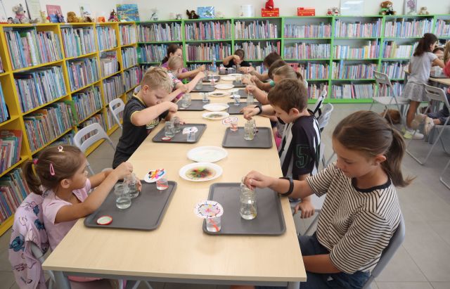Na zdjęciu znajduje się znajduje się około 12 dzieci przy złączonych stolikach i mieszają wodę w słoikach, na stolikach przed nimi leżą talerzyki z cukierkami, które puszczają kolor we wodzie. W tle znajdują się półki z książkami.