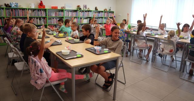 Na zdjęciu znajdują się dzieci przy złączonych w dwóch rzędach stolikach i mają podniesione do góry ręce. Na stolikach przed nimi leżą tacki z miseczkami i szklanką. W tle znajdują się półki z książami i okna.