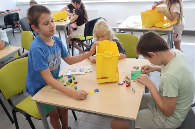 Na zdjęciu znajdują się dzieci przy czterech stolikach i układają klocki LEGO.