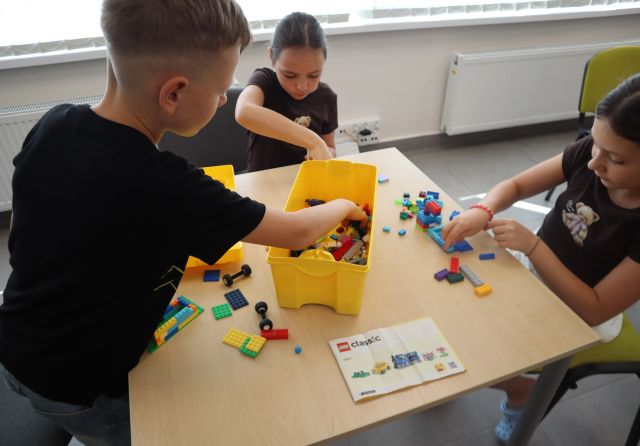 Na zdjęciu znajdują się dwie dziewczynki siedzące przy stoliku i jeden chłopiec i budują swoje światy z klocków LEGO. W tle znajdują się okna.