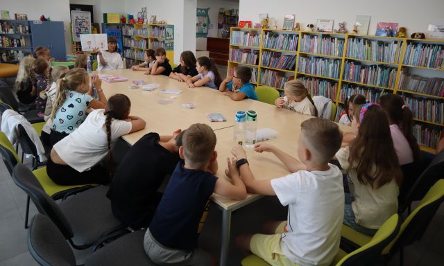 Na zdjęciu znajduje się 20 dzieci przy złączonych stołach, wraz z nimi siedzi kobieta, która pokazuje im ilustracje w książce. Na stolikach leżą koraliki do bransoletek w pudełeczkach. W tle znajdują się półki z książkami.