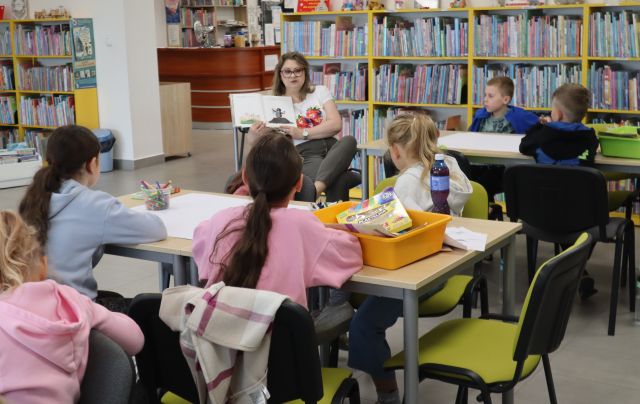 Na zdjęciu znajdują się dzieci przy złączonych stolikach w dwóch rzędach, na których leżą przybory potrzebne do pracy. Pomiędzy nimi na krześle siedzi kobieta i pokazuje im ilustracje w książce. W tle znajdują się półki z książkami.