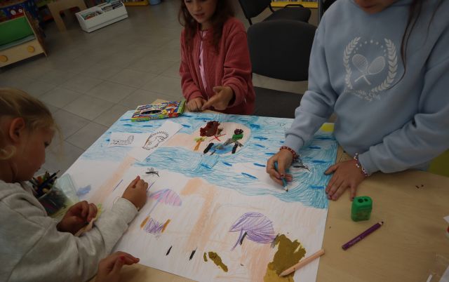 Na zdjęciu znajdują się trzy dziewczynki przy stoliku i razem kolorują wykonane przez siebie ilustracje na dużej kartce.
