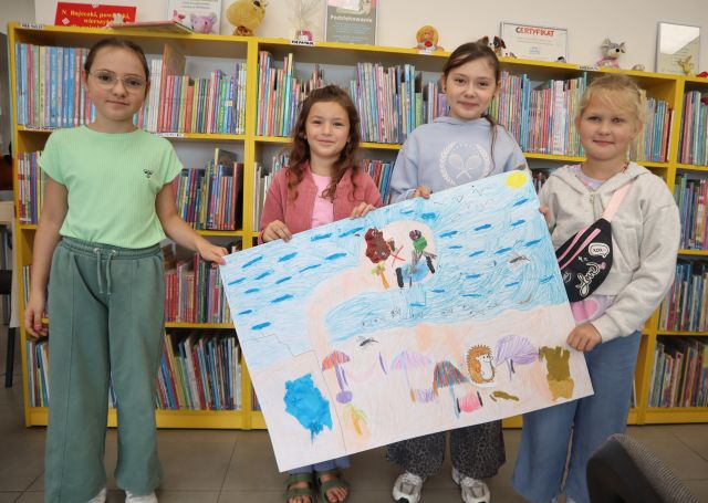 Na zdjęciu znajdują się cztery dziewczynki trzymająca wykonaną przez siebie ilustrację na dużej kartce przedstawiającą Krecika na wyspie przy morzu. W tle znajdują się półki z książkami.