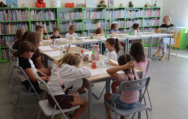 Na zdjęciu znajdują się dzieci przy złączonych w literę L stolikach, na brzegu stołów siedzi kobieta i czyta książkę. W tle znajdują się półki z książkami.