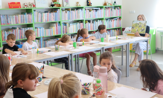 Na zdjęciu znajdują się dzieci przy złączonych stolikach, na brzegu siedzi kobieta i pokazuje dzieciom ilustrację w książce. Stoliki są owinięte folią, leżą na nich kredki, kartki. W tle znajdują się półki z książkami.