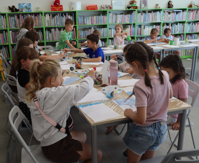 Na zdjęciu znajdują się dzieci przy złączonych stolikach i malują na kartkach wspomnienia z wakacji. W tle znajdują się półki z książkami.