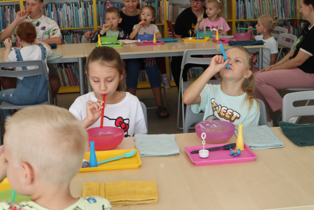 Na zdjęciu w centrum znajdują się dwie dziewczynki robiące bańki mydlane przy złączonych stolikach, przed nimi znajdują się miseczki, tacki oraz ręczniki. W tle znajduje się drugi rząd stolików z dziećmi, które równie puszczają bańki mydlane, a za nimi znajdują się półki z książkami.