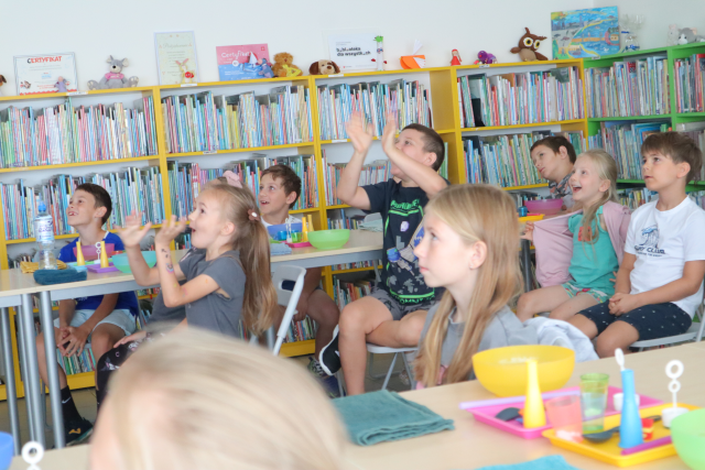 Na zdjęciu znajdują się dzieci przy złączonych w dwóch rzędach stolikach, dwójka dzieci ma podniesione ręce do góry, na stolikach leżą miseczki, tacki. W tle znajdują się półki z książkami.