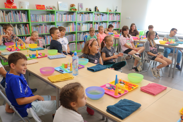 Na zdjęciu znajdują się dzieci przy stolikach w dwóch rzędach. Na stolikach znajdują się miseczki, ręczniki, tacki z akcesoriami. W tle znajdują się półki z książkami i okna.