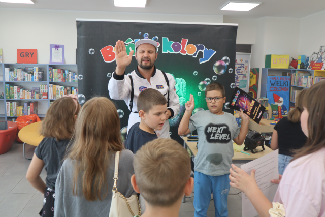 Na zdjęciu znajduje się piątka dzieci, jeden z nich trzyma dyplom, a obok niego stoi mężczyzna z podniesioną ręką. W tle znajduje się czarny baner z napisem, a w tle półki z grami.
