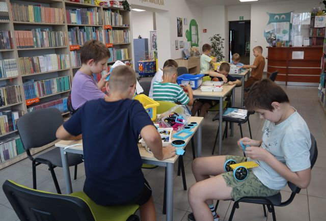 Na zdjęciu znajduje się dziewięć chłopców przy trzech stolikach, przy których układają klocki LEGO. W tle znajdują się półki z książkami oraz biurko biblioteczne.