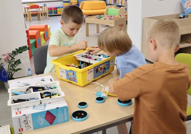 Na zdjęciu znajduje się trzech chłopców przy stoliku, dwóch wspólnie szuka odpowiednich klocków w pojemniku, a jeden składa klocki. W tle znajduje się pomieszczenie biblioteczne ze stolikiem do zabawy, pufami.