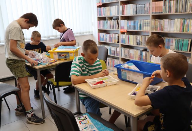 Na zdjęciu znajduje się 6 chłopców przy dwóch stolikach, w trójkach układają zestawy LEGO znajdujące się na stolikach przed nimi. W tle znajdują się półki z książkami oraz okna.