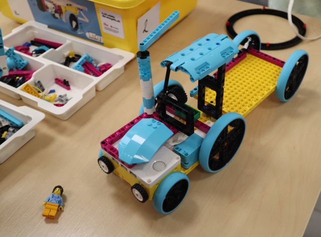 Na zdjęciu znajduje się zbudowany z klocków LEGO ciągnik z przyczepą, na stole dookoła leżą klocki w plastikowych pudełeczkach.