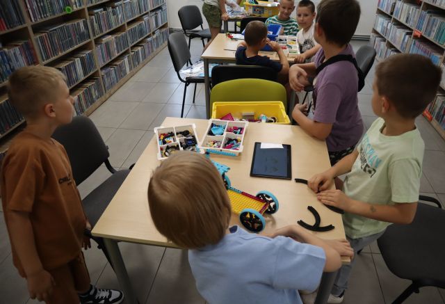 Na zdjęciu znajduje się 7 chłopców przy dwóch stolikach i układają znajdujące się przed nimi klocki LEGO, na pierwszym stoliku leży tablet, na którym chłopcy kodują swojego robota. W tle znajdują się półki z książkami.