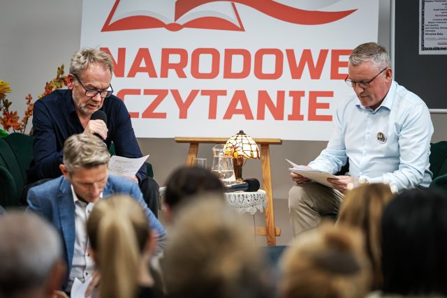 Na zdjęciu znajduje się dwóch mężczyzn na scenie, którzy czytają, pomiędzy nimi znajduje się stolik z lampką oraz biała tablica z napisem Narodowe Czytanie. Przed sceną na fotelu siedzi mężczyzna a przed nimi widownia.