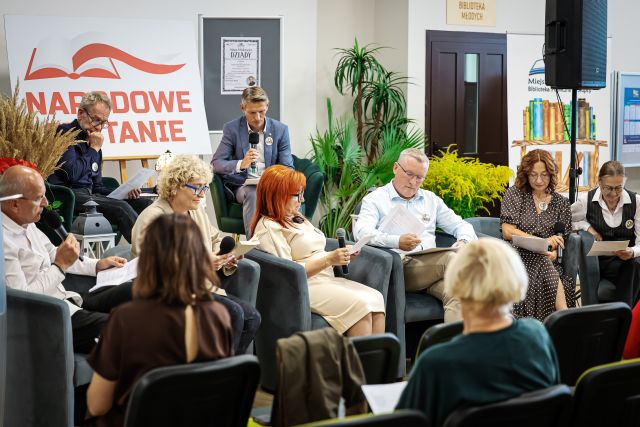 Nazdjęciu znajdują się dwie osoby siedzące na scenie oraz sześć przed sceną. Na scenie znajduje się stolik z lampką oraz biała tablica z czerwonym napisem Narodowe Czytanie, przy scenie stoją kwiaty oraz baner z książkami i głośnik. Przed czytającymi znajduje się widownia.
