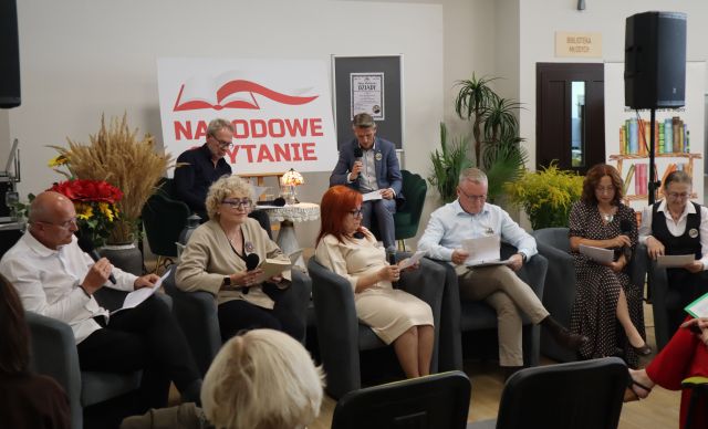 Na zdjęciu znajduje się osiem osób, dwóch mężczyzn na scenie oraz sześć osób przed sceną na fotelach. Na scenie znajduje się stolik z lampką, tablica z napisem Narodowe Czytanie oraz plakat promujący to wydarzenie, obok sceny znajdują się kwiaty, drzwi, baner z książkami oraz głośnik na statywie.