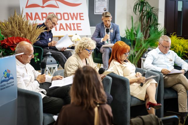 Na zdjęciu znajduje się dwóch mężczyzn siedzących na scenie oraz cztery osoby siedzące na fotelach przed sceną. Na scenie w tle znajdują się kwiaty oraz biała tablica z czerwonym napisem Narodowe Czytanie.