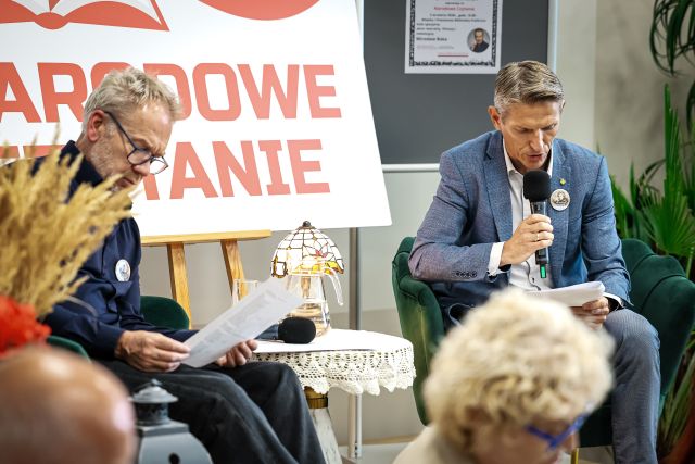 Na zdjęciu znajduje się dwójka mężczyzn na scenie z mikrofonami oraz kartkami w ręku, pomiędzy nimi znajduje się stolik z lampką. Na scenie znajdują się kwiaty, biała tablica z czerwonym napisem Narodowe Czytanie oraz plakat promujący to wydarzenie.