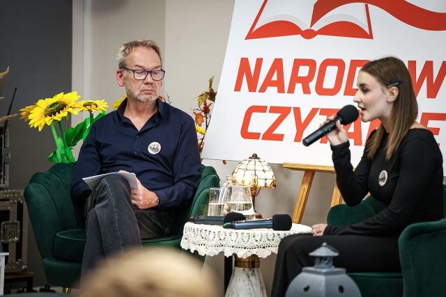 Na zdjęciu znajdują się dwie osoby siedzące na fotelach na scenie, mężczyzna oraz dziewczyna, która śpiewa. Pomiędzy nimi znajduje się stolik z wodą oraz lampką, a w tle znajdują sie kwiaty oraz biała tablica z czerwonym napisem Narodowe Czytanie.