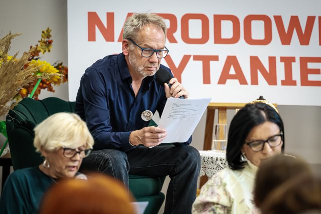 Na zdjęciu znajduje się siedzący na scenie mężczyzna z mikrofonem oraz kartką, przed sceną siedzą dwie kobiety. W tle znajduje się biała tablica z czerwonym napisem napisem Narodowe Czytanie.
