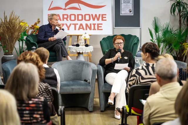 Na zdjęciu znajduje się widownia, po lewej i prawej stronie, a przed nimi na fotelu siedzi kobieta i mówi przez mikrofon, obok niej stoją puste fotele. Na scenie za kobietą siedzi mężczyzna, który trzyma w ręce mikrofon oraz kartki, drugi fotel jest wolny. Na scenie za mężczyzną znajdują się kwiaty, stolik z lampką i wodą, tablica z napisem Narodowe Czytanie oraz plakat promujący to wydarzenie.